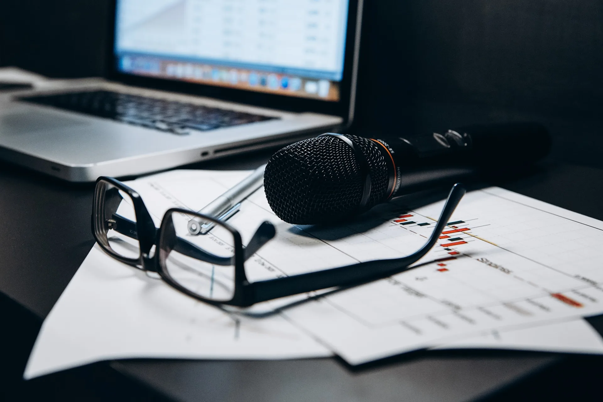 Microphone, glasses and charts on an office desk next to a laptop
