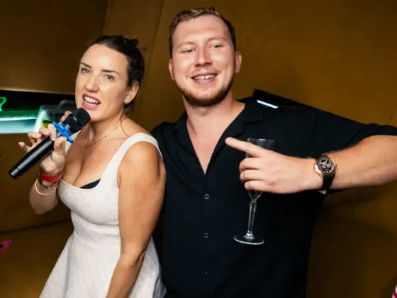 Chica con vestido blanco canta al micrófono junto a un chico sonriente con una copa en una sala de karaoke