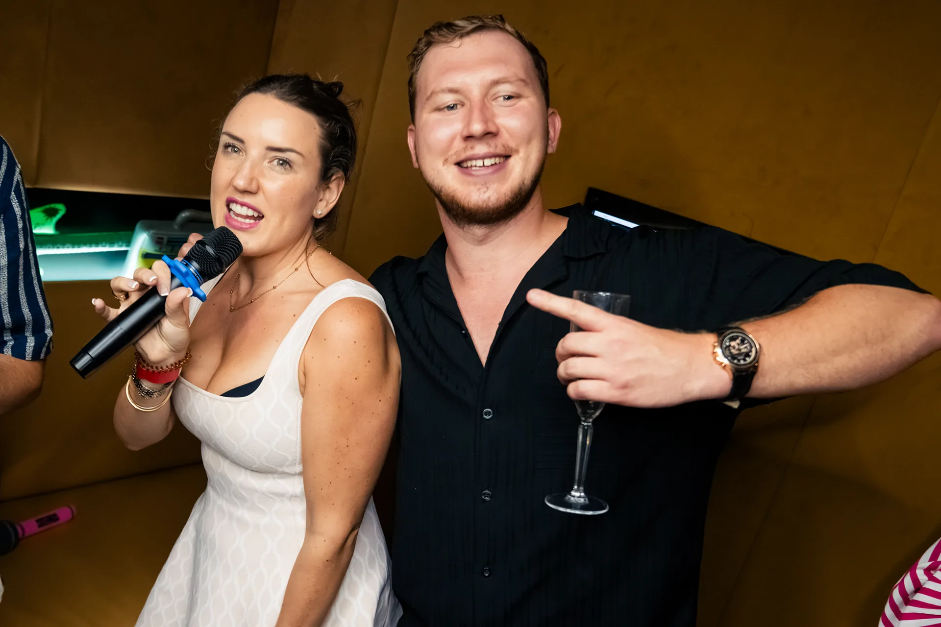 Woman in a white dress singing into a microphone next to a smiling man with a glass in a karaoke room