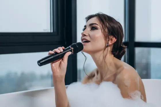 Chica cantando con un micrófono inalámbrico en una bañera con espuma junto a la ventana