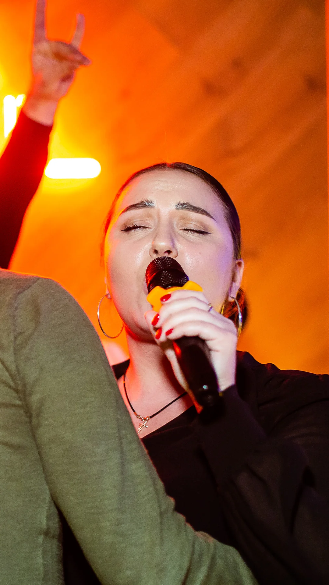 Woman singing into a microphone at a karaoke party as guests raise rock horns in front of a neon sign