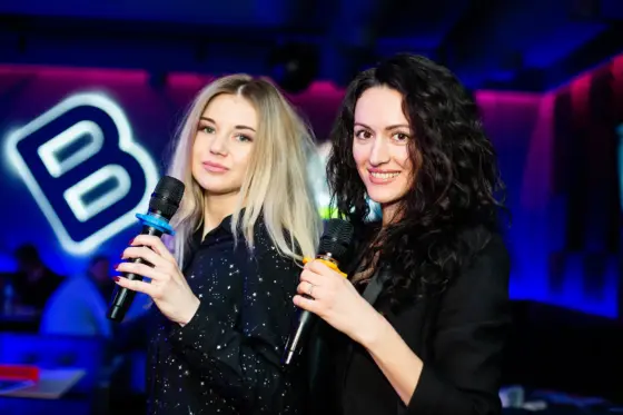 Dos chicas con micrófonos de karaoke sonríen en una fiesta con luces de neón