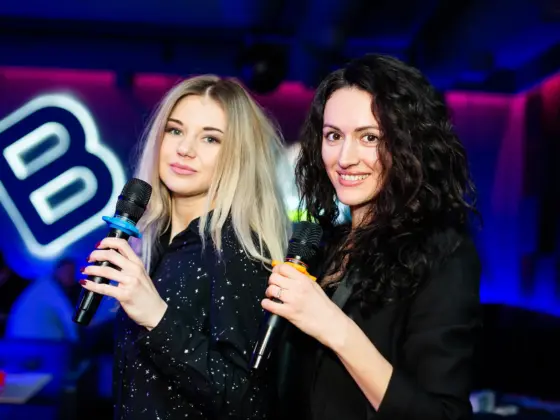 Two women holding karaoke microphones smiling at a party in neon light