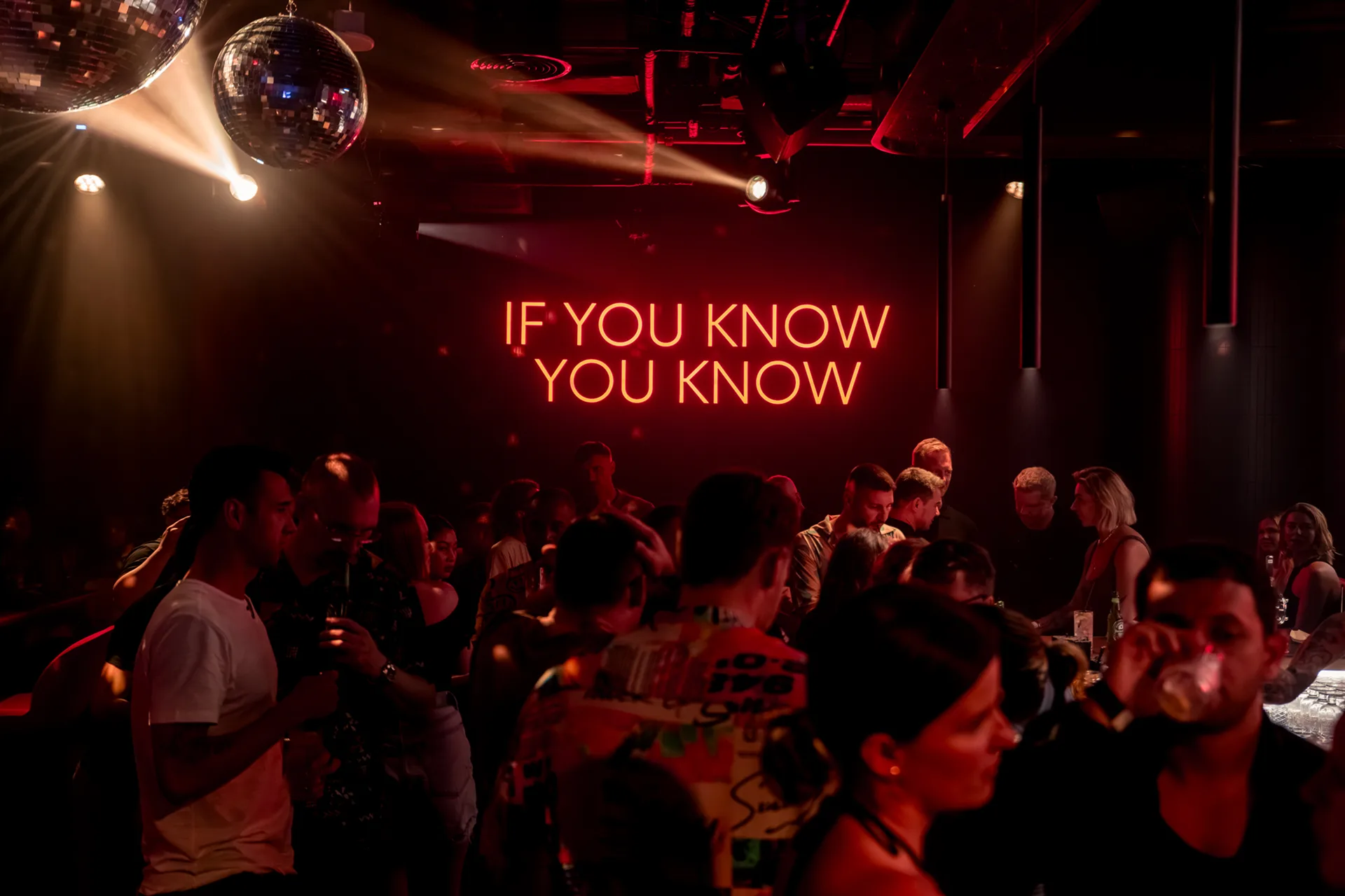 Neon sign reading “If you know you know” above a crowd of guests in a karaoke club with disco balls