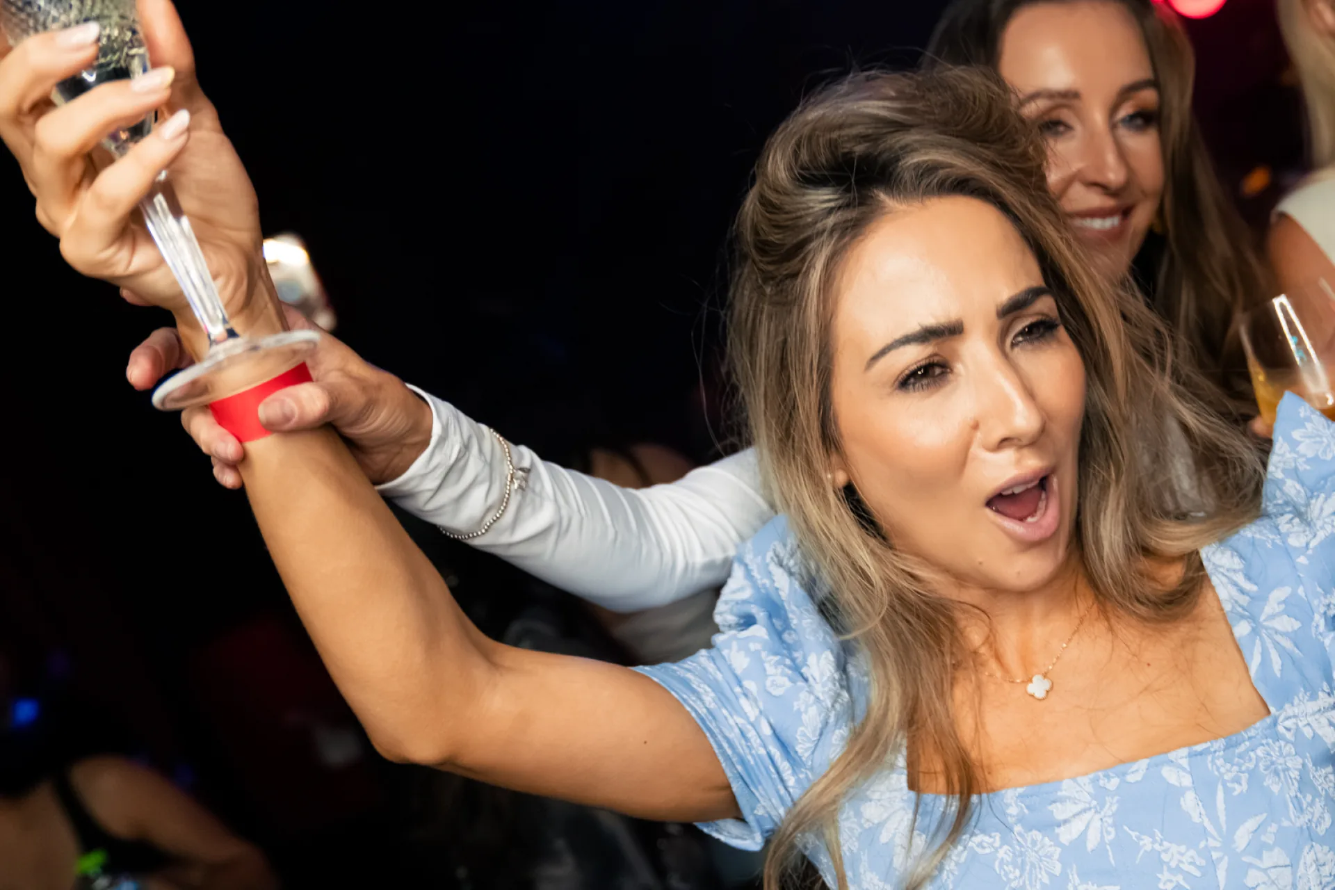 A woman in a light blue dress singing and raising a glass with friends at a karaoke club party