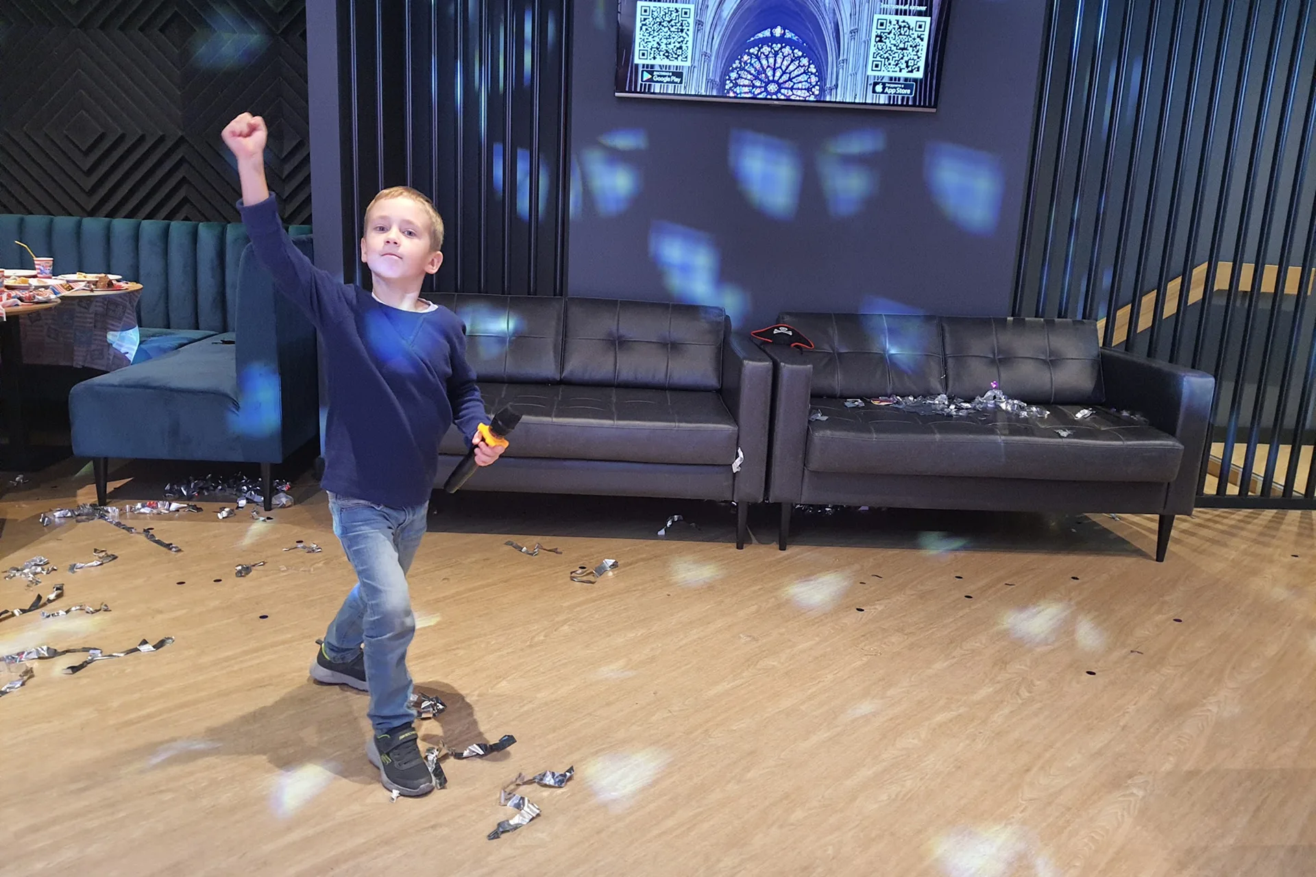 A boy with a microphone happily raising his fist at a kids’ party in a karaoke room with confetti on the floor