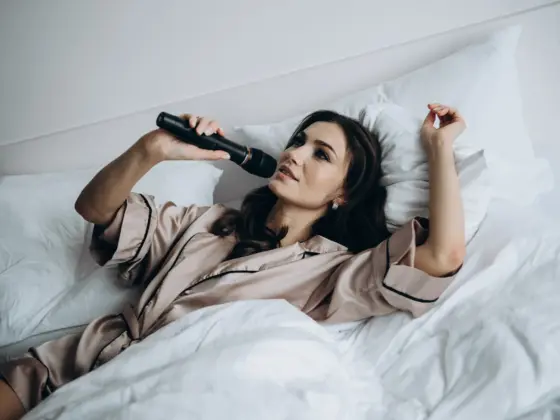 Young woman in silk pajamas lying in bed and singing into a microphone