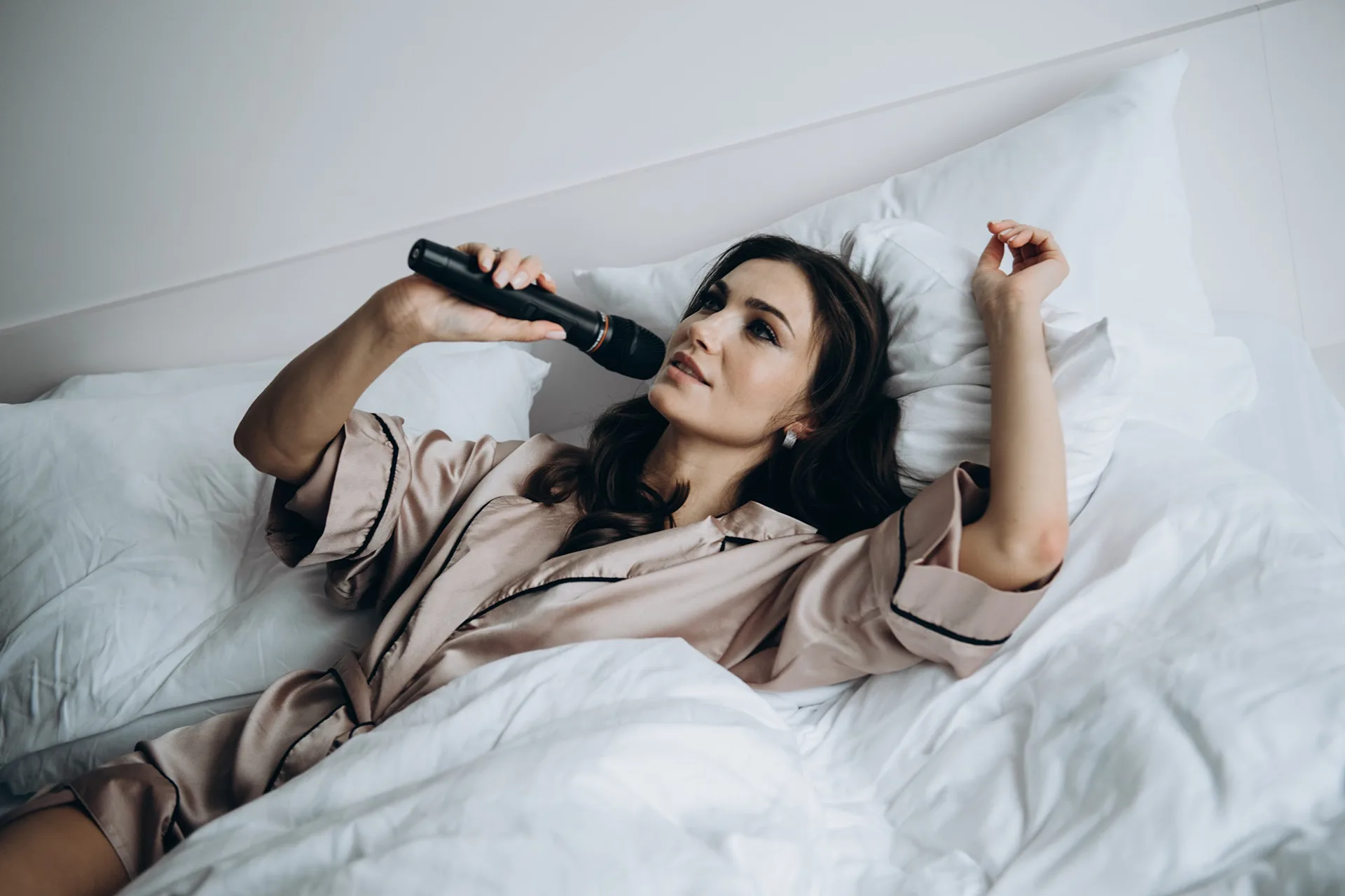 Young woman in silk pajamas lying in bed and singing into a microphone