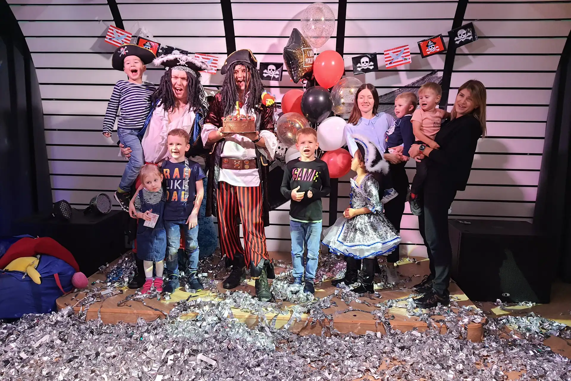 Kids and parents with pirate entertainers and a cake on stage at a children’s party among confetti