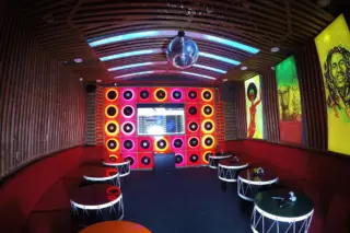 Karaoke room interior: a wall of glowing vinyl records, a screen, a disco ball, red sofas and drum-shaped tables