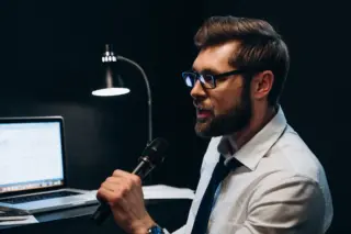 A man in glasses, a shirt and tie singing into a microphone at an office desk with a laptop