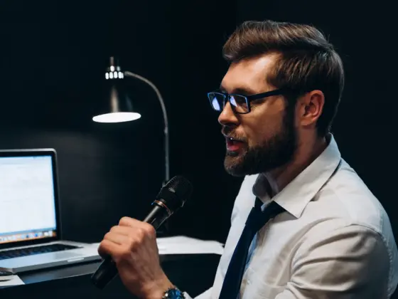 A man in glasses, a shirt and tie singing into a microphone at an office desk with a laptop