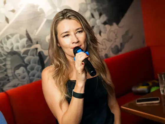 A woman with long blonde hair singing into a microphone on a red sofa in a karaoke bar