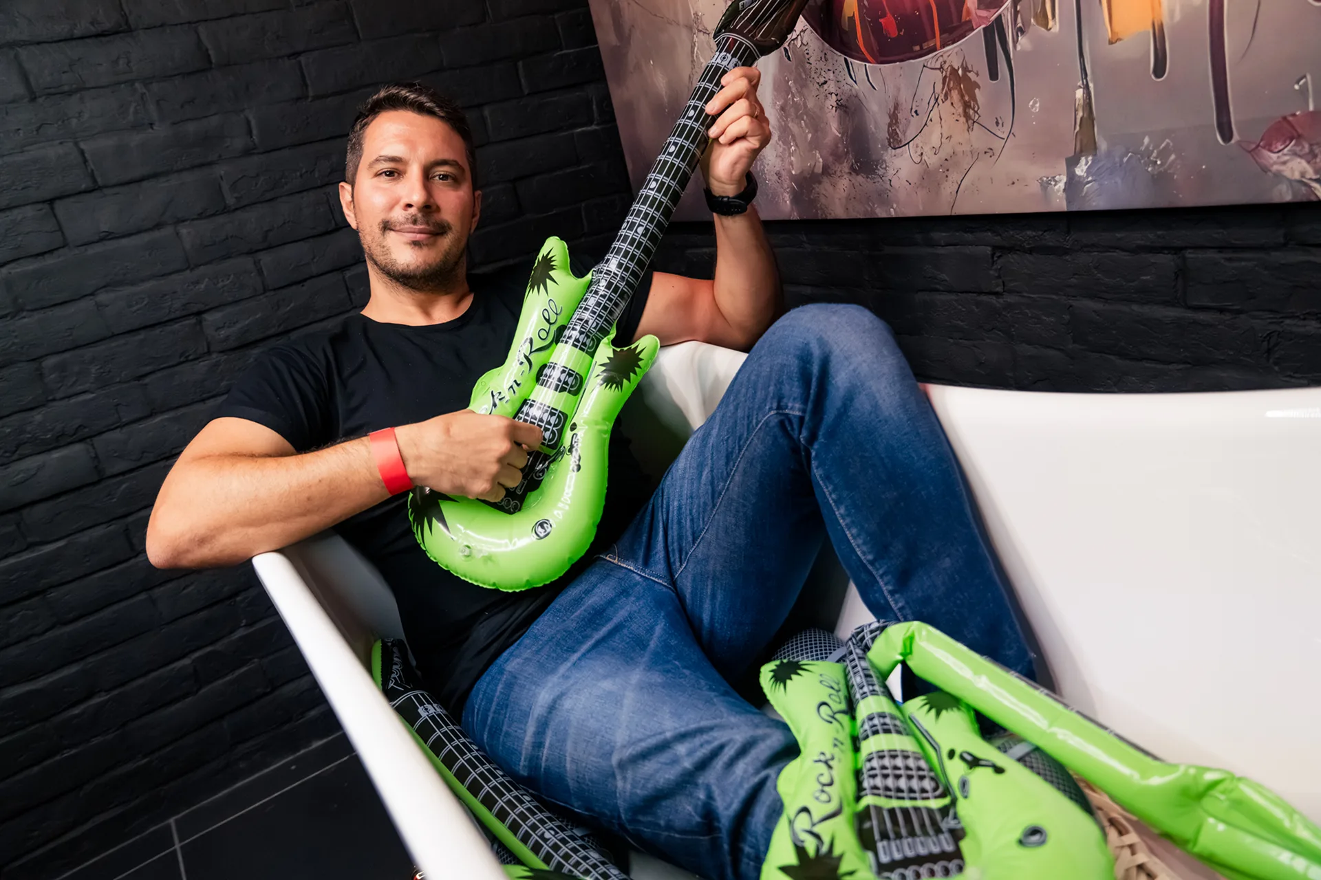 A smiling man sitting in a white armchair holding a green inflatable guitar
